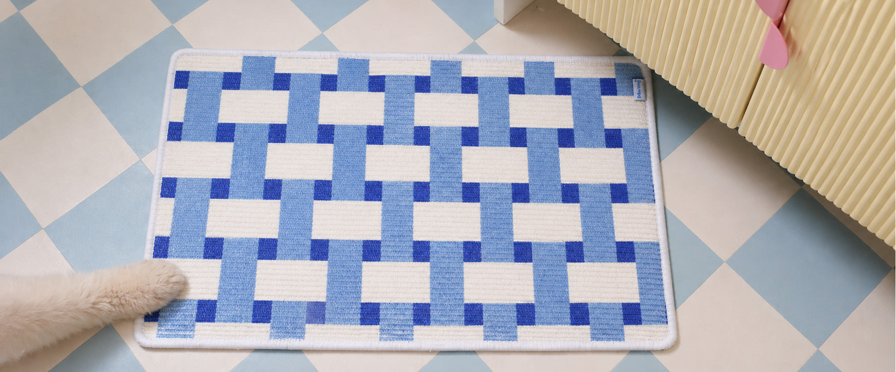 Grid Play scratching pad on a chequered floor, a cat's paw entering the frame